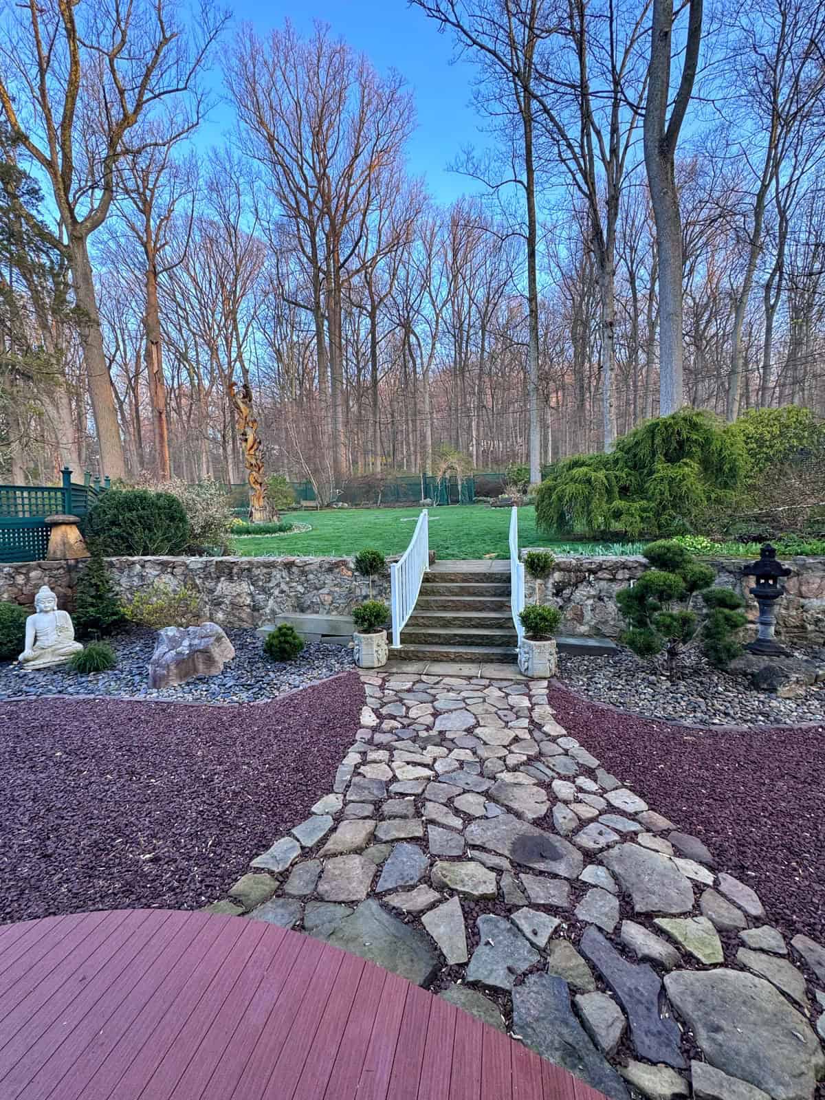 A beautiful backyard zen garden that has a stone path leads from a wooden deck to stairs with white railings, surrounded by garden statues, rocks, shrubs, and trees. In the background, there is a grassy lawn and a forest of leafless trees. 