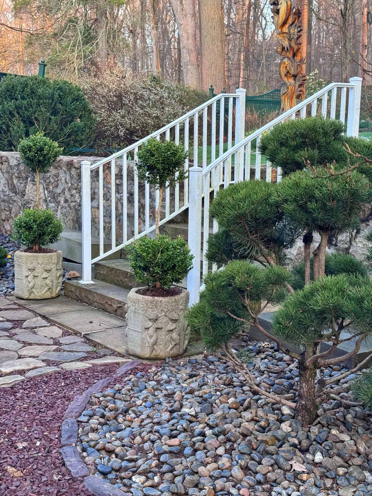 A beautiful backyard zen garden with a white metal railing staircase, potted topiary plants, a stone path, small rocks, and trimmed shrubs. Trees and a stone wall are in the background.