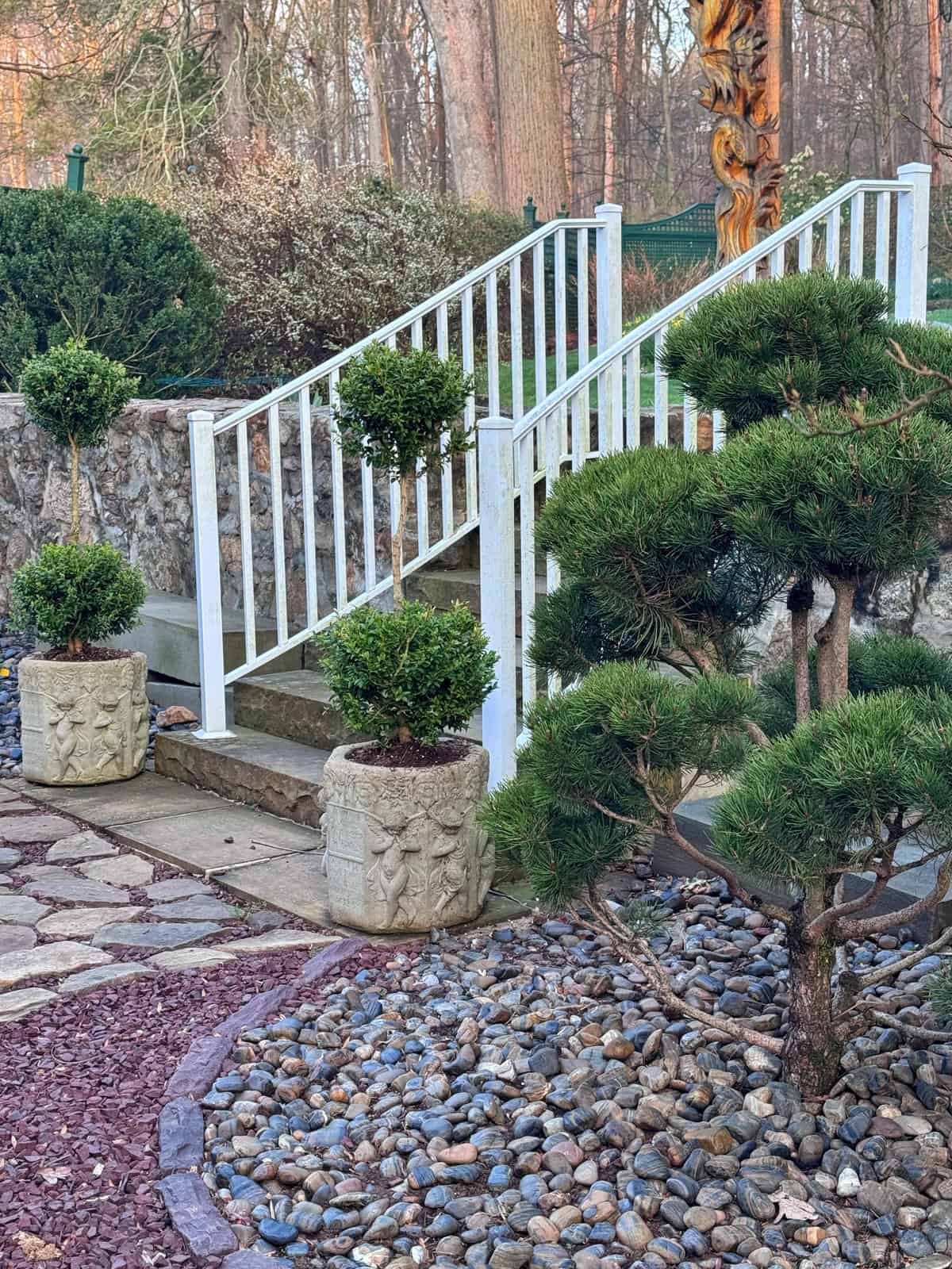 White outdoor stairs with a metal railing lead up beside stone planters containing shaped topiary shrubs. Decorative rocks, a pruned bush, and paving stones are arranged along the walkway. Trees are visible in the background of a beautifully landscaped backyard zen garden.