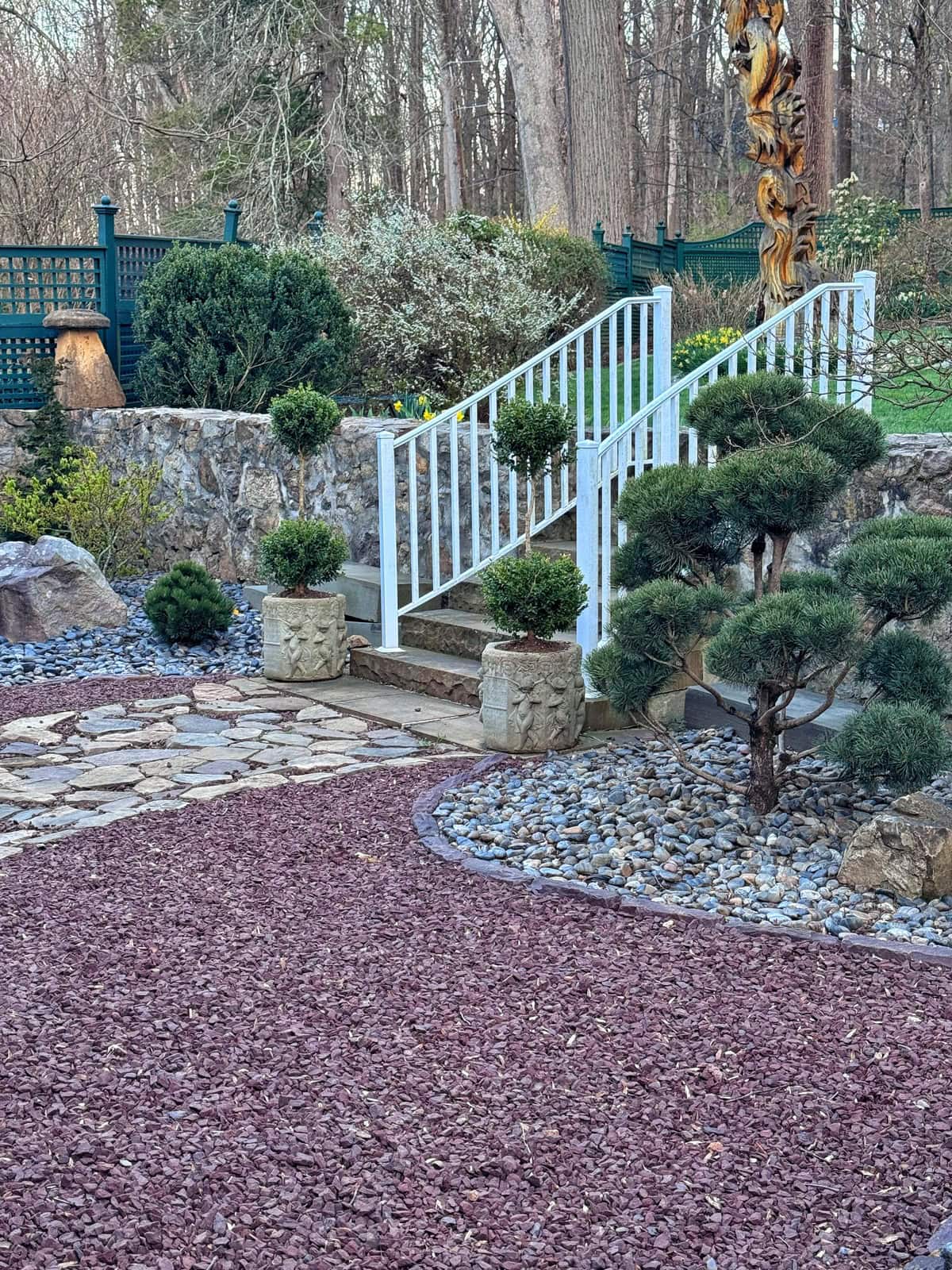 A landscaped zen garden features a stone pathway, red gravel, decorative shrubs, and a white railing staircase leading to a higher level. Rocks, small bushes, and trees complete the tranquil scene in the background.