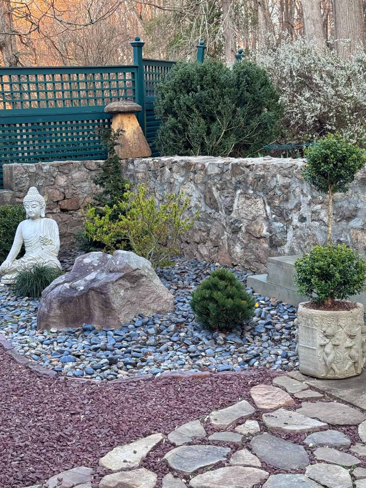A serene backyard zen garden features a Buddha statue, a large rock, small shrubs, and a stone planter on a bed of blue pebbles, bordered by a stone path, a rock wall, and green lattice fencing in the background.