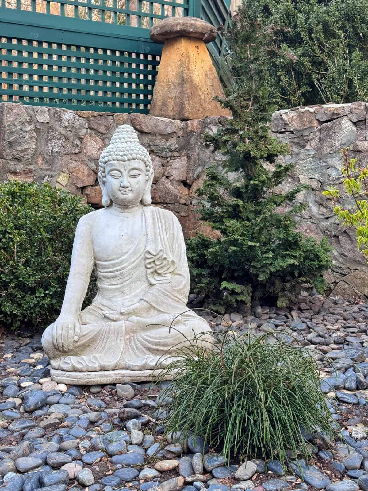 A white stone Buddha statue sits in a meditative pose on a bed of pebbles, surrounded by green shrubs and a small tree, with a stone wall and lattice fence in the background of a beautifully landscaped zen garden