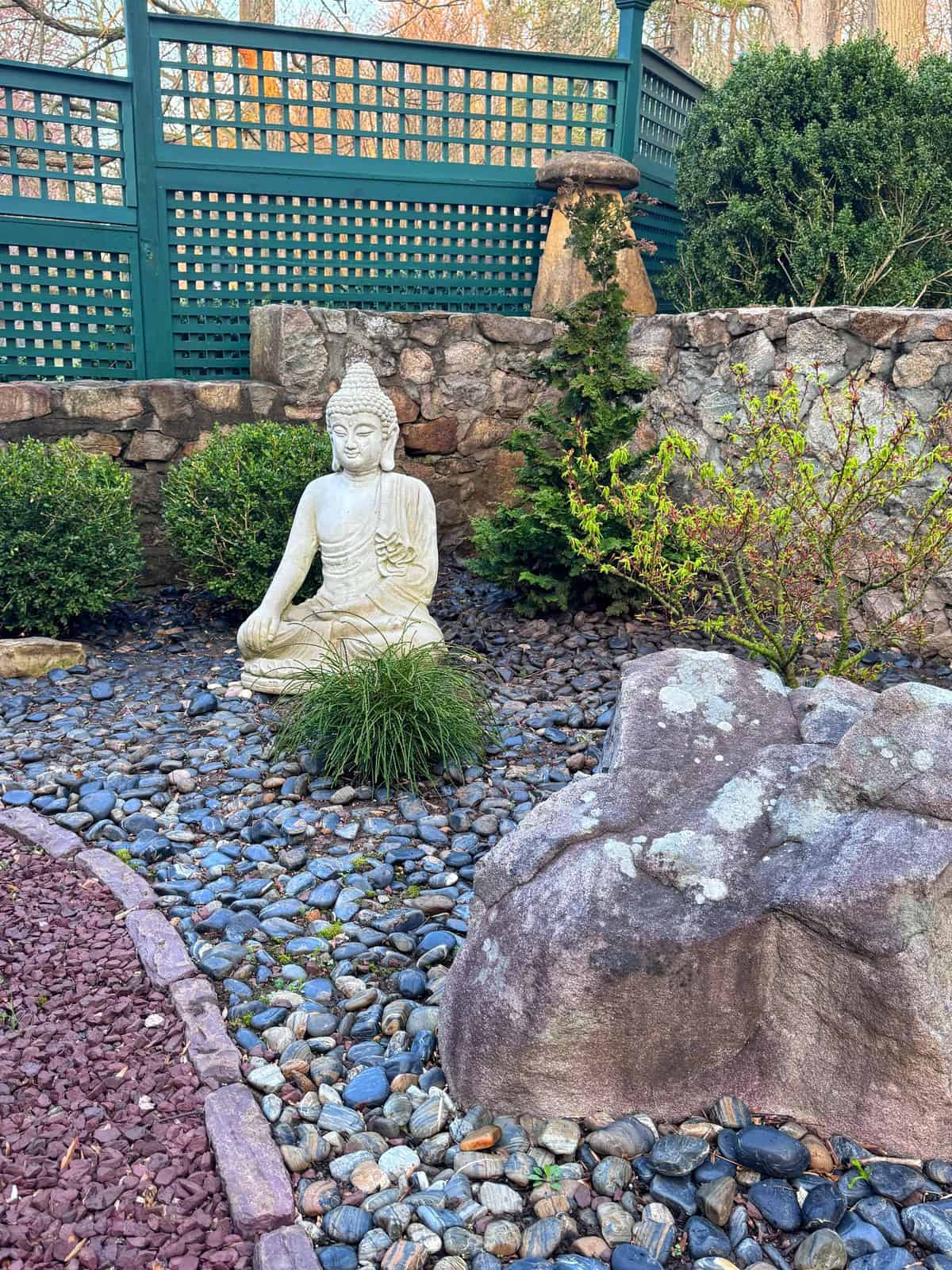 A white Buddha statue sits on gray pebbles in a serene zen garden, surrounded by green bushes, rocks, and a stone wall with a green lattice fence in the background.