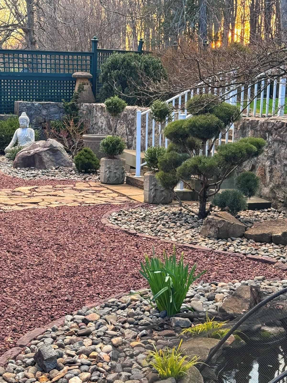 A serene zen garden with a gravel path, rocks, trimmed bushes, a pond, and a Buddha statue. Steps and a white railing lead up to a fence, with the sun setting through trees in the background.