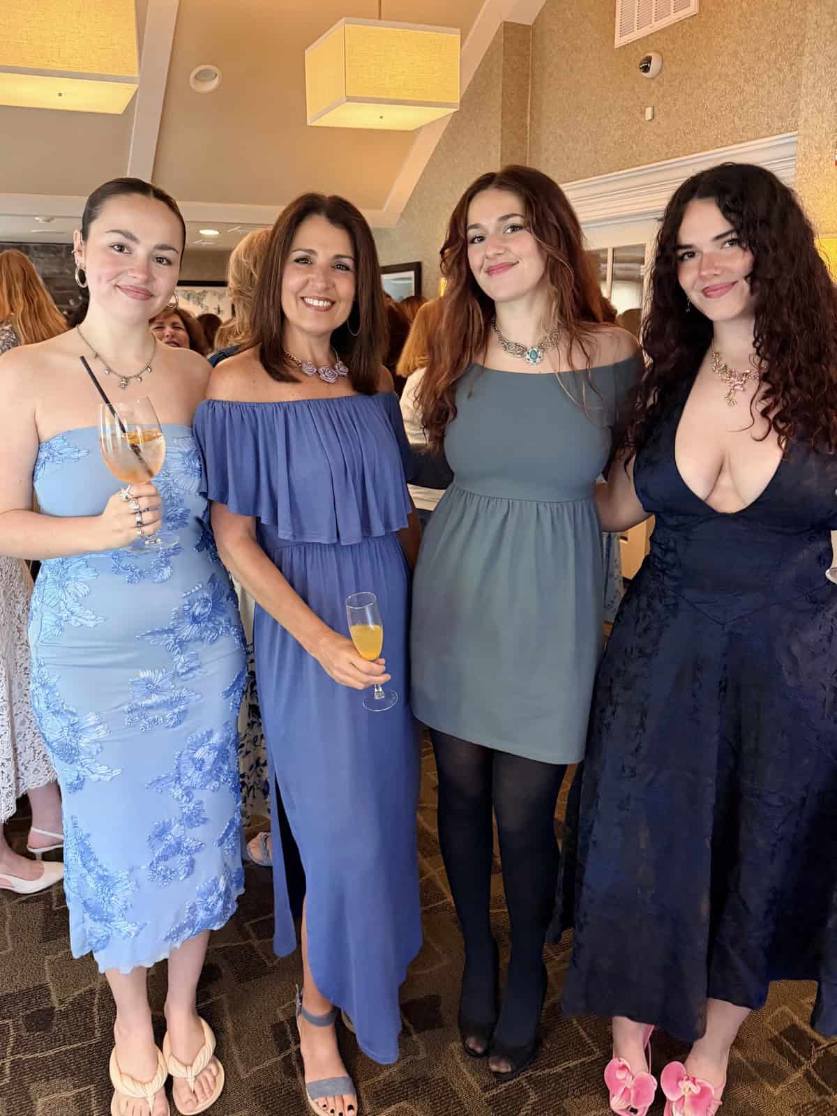 Four women (stacy ling and her daughters) stand together at an indoor event, dressed in blue and navy dresses, holding glasses and smiling at the camera. The background shows other guests and soft lighting fixtures.