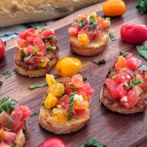 A wooden board with toasted baguette slices topped with chopped tomatoes, basil, and garlic. Whole grape tomatoes and basil leaves are scattered around, with a baguette loaf in the background.