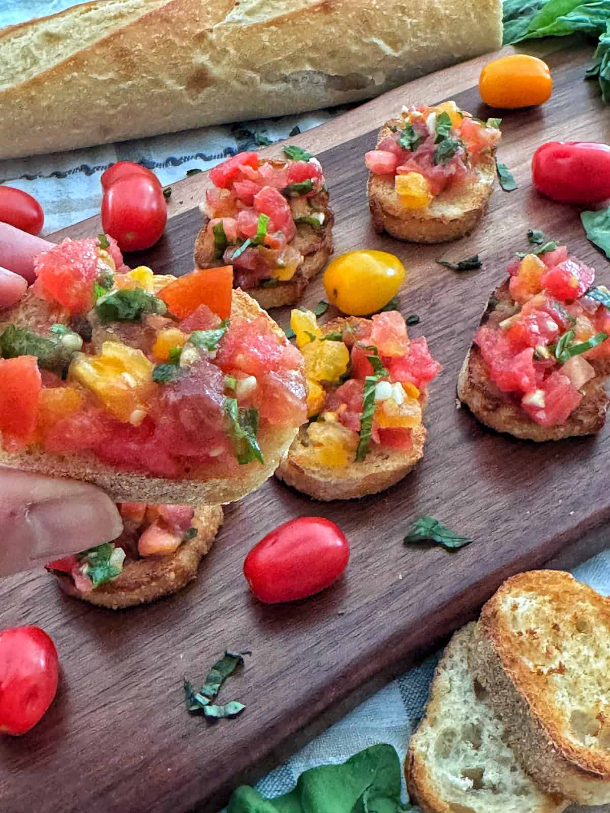 A hand holds a piece of bruschetta topped with diced tomatoes and herbs. More bruschetta, grape tomatoes, and fresh basil are arranged on a wooden board, with a baguette in the background.