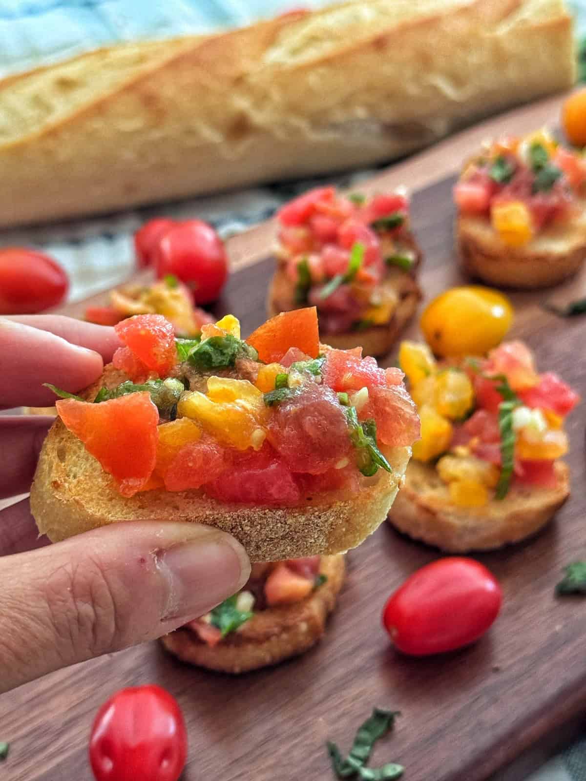 A hand holds a piece of bruschetta topped with diced tomatoes, basil, and other vegetables. More bruschetta pieces, cherry tomatoes, and a baguette are visible on a wooden board in the background.