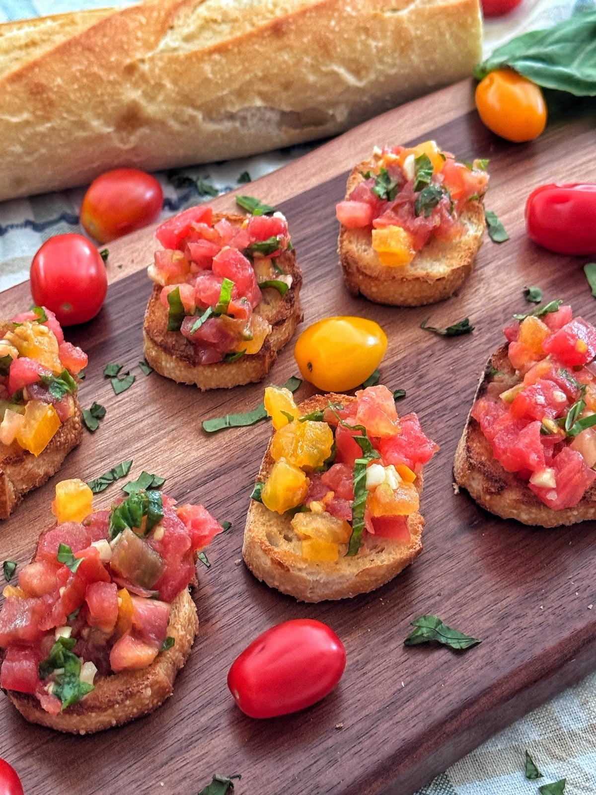 A wooden board with several slices of toasted bread topped with diced red and yellow tomatoes, chopped herbs, and surrounded by grape tomatoes and a baguette.