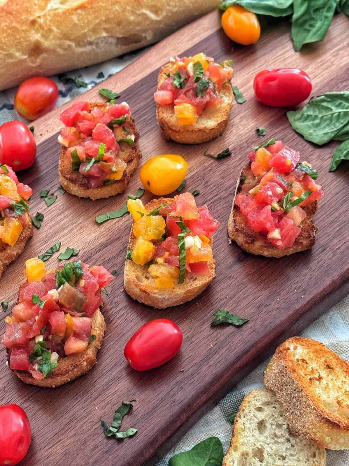 Slices of toasted bread topped with diced red and yellow tomatoes, fresh basil, and herbs are arranged on a wooden board, surrounded by grape tomatoes, basil leaves, and a loaf of bread in the background.