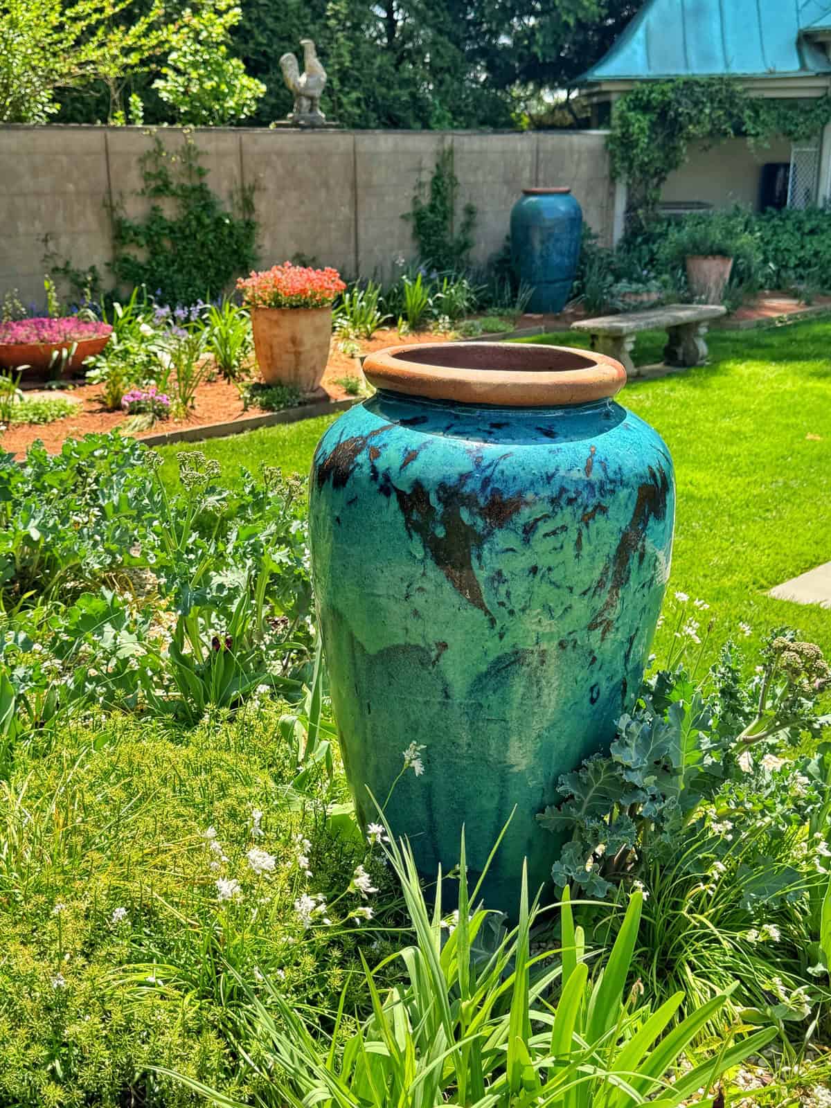 A large, turquoise ceramic pot with a rustic, speckled finish sits in a garden, surrounded by lush green plants and white flowers.