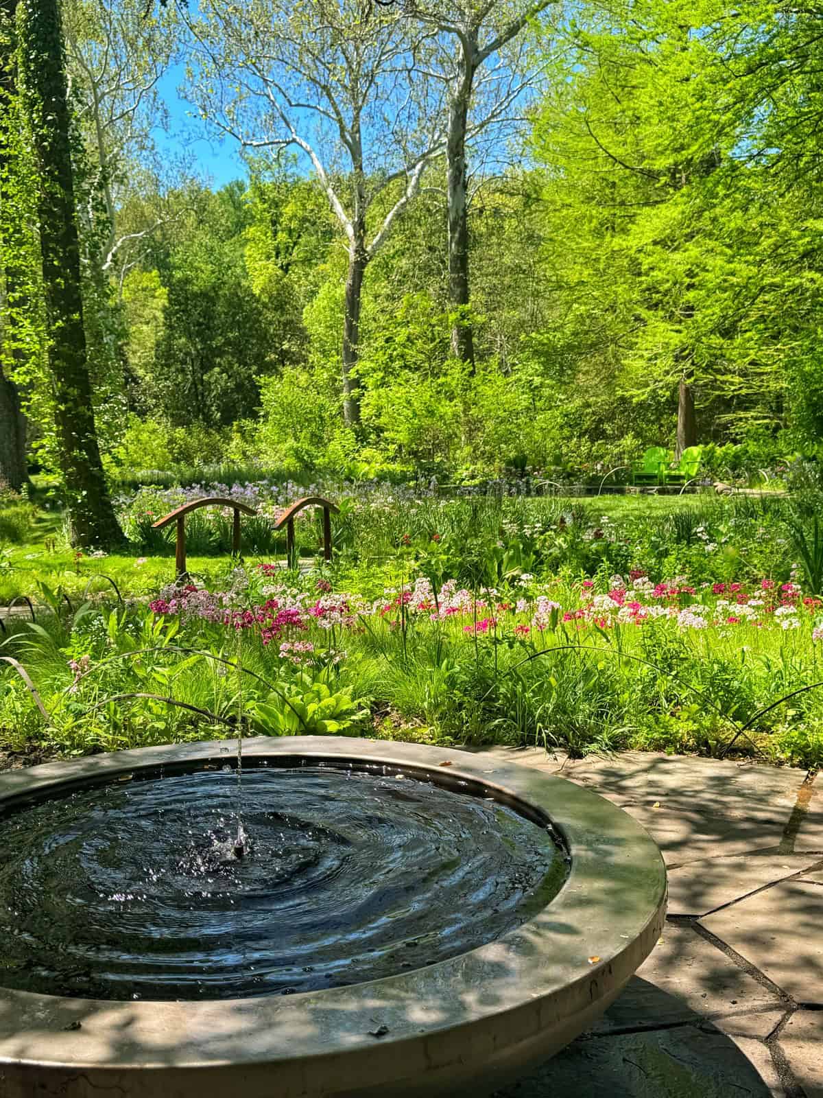 A circular fountain with rippling water in the foreground, leading to a lush garden with colorful flowers, small arched bridges, and mature trees under a clear blue sky.