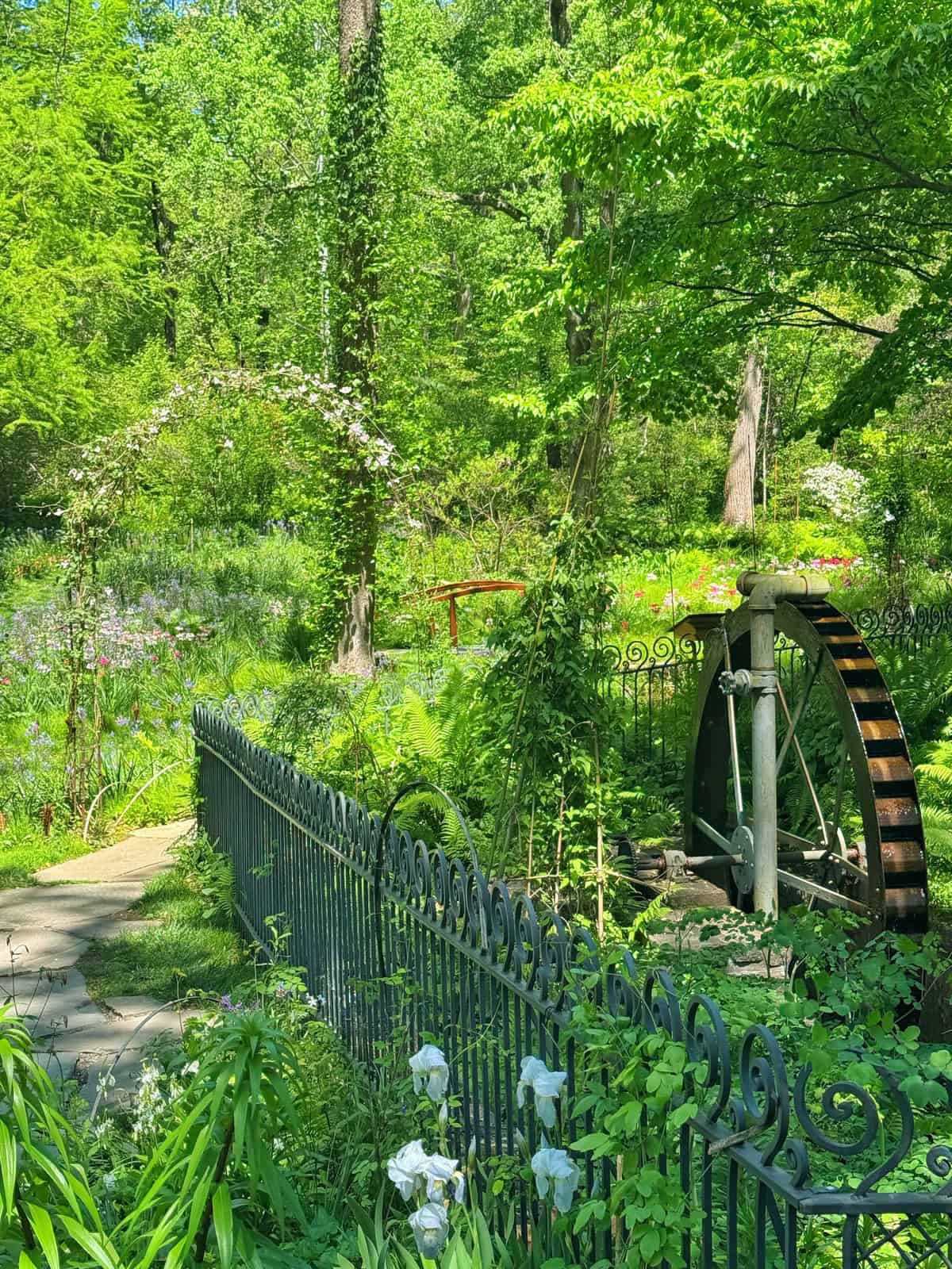 A weathered water wheel sits behind a decorative iron fence in a lush, sun-dappled garden. Wildflowers and greenery abound under a canopy of trees.