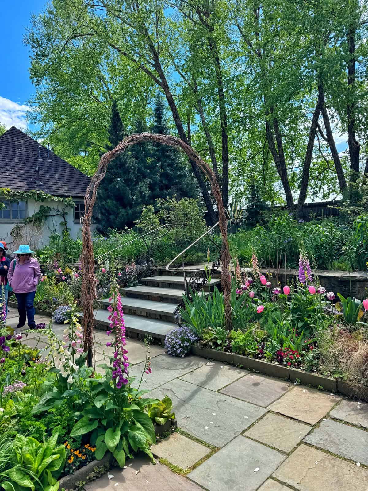A rustic archway frames a garden path and stone steps leading to a house surrounded by lush greenery and blooming flowers like foxgloves and tulips.