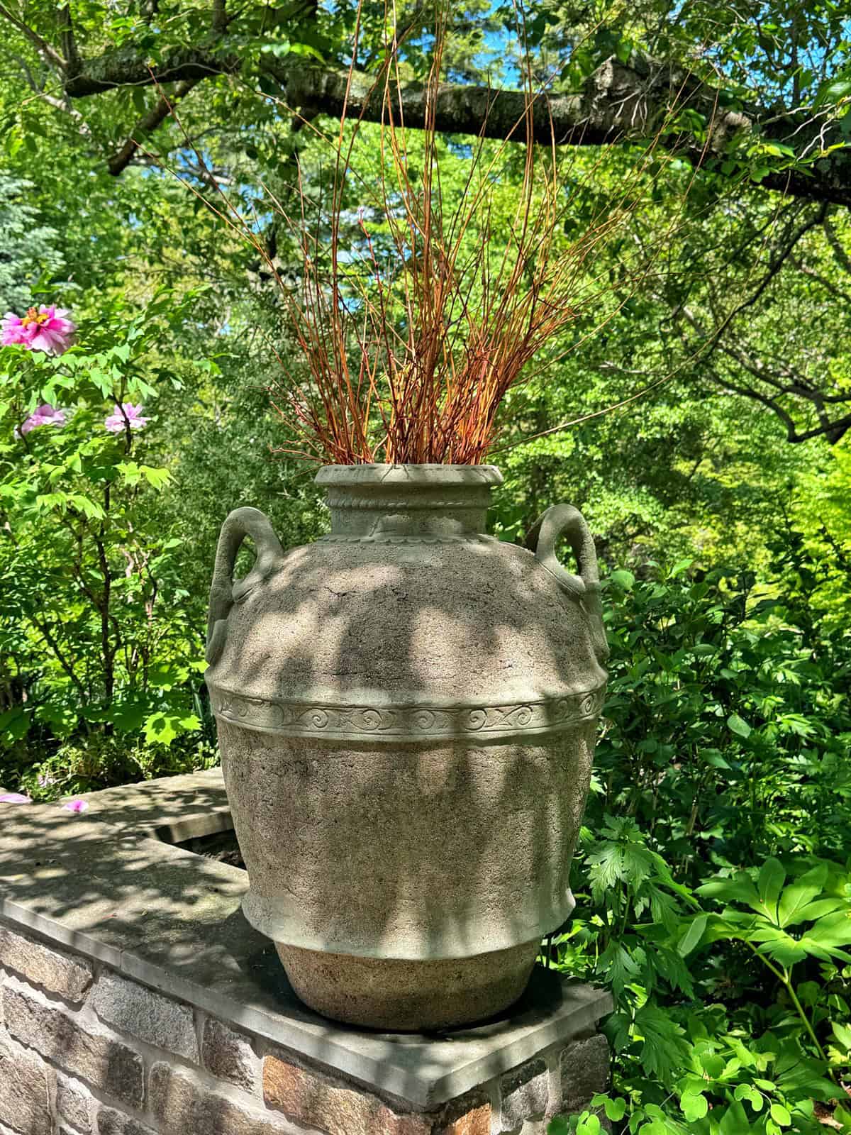 A large, textured, urn-shaped planter with two handles, filled with reddish-brown twigs, sits on a stone wall surrounded by lush green foliage and pink peonies.