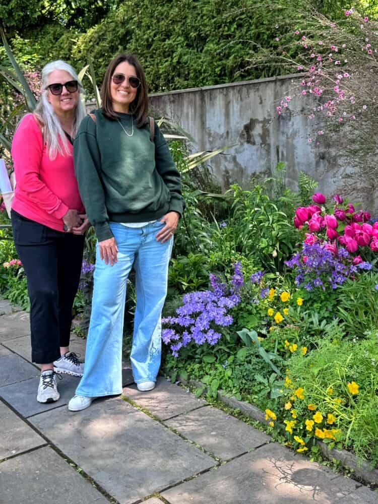 Two women smile while standing on a stone path next to a vibrant garden with pink, purple, and yellow flowers. Both wear sunglasses; one is in a pink top and black pants, the other in a green sweatshirt and light blue jeans.