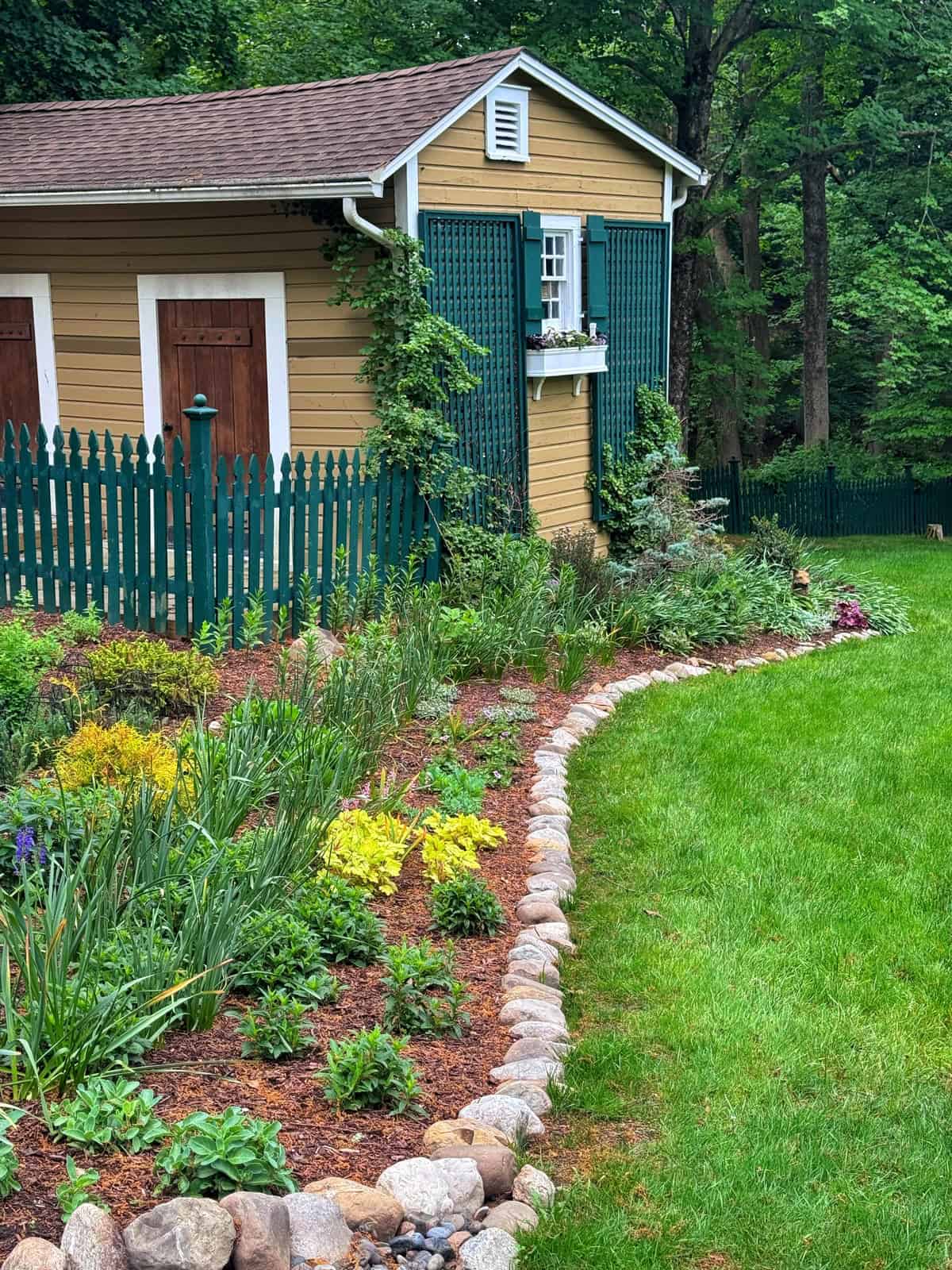 A yellow shed with green trim and a small window sits by a green picket fence. A landscaped garden with rocks, flowers, and plants lines a neatly mowed lawn, bordered by trees in the background.