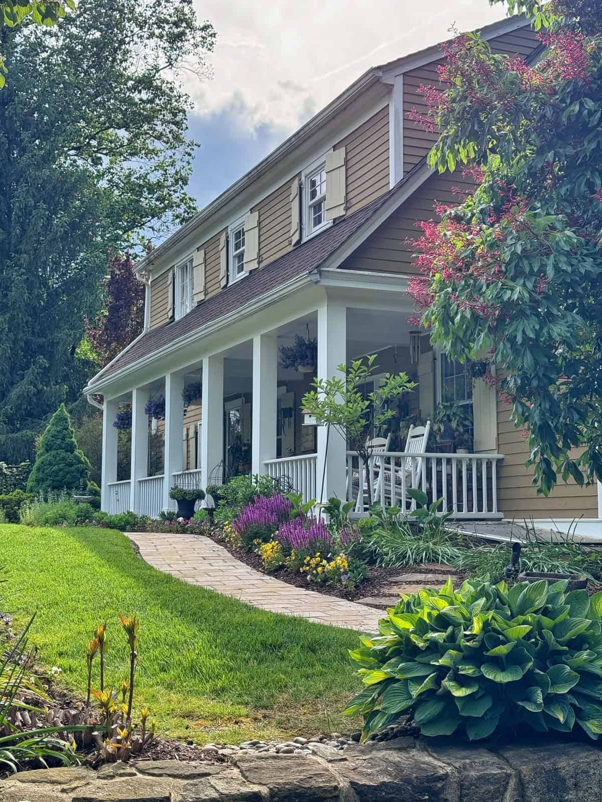 A two-story tan house with white trim, a large front porch with rocking chairs, hanging flower baskets, and lush landscaping featuring green grass, colorful flowers, and shrubs along a stone walkway.