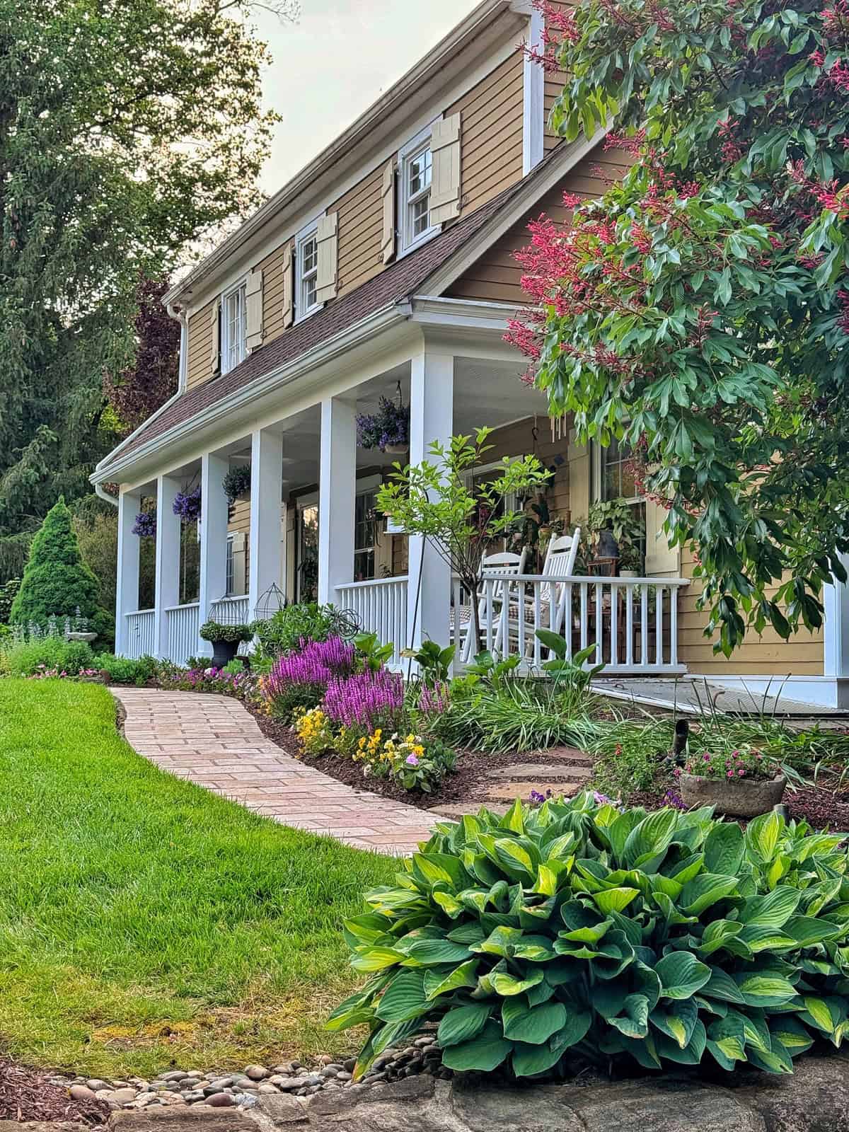A charming house with tan siding, white trim, and a large front porch. The porch has rocking chairs and hanging flower baskets. The yard features lush green plants, blooming flowers, and a brick walkway.