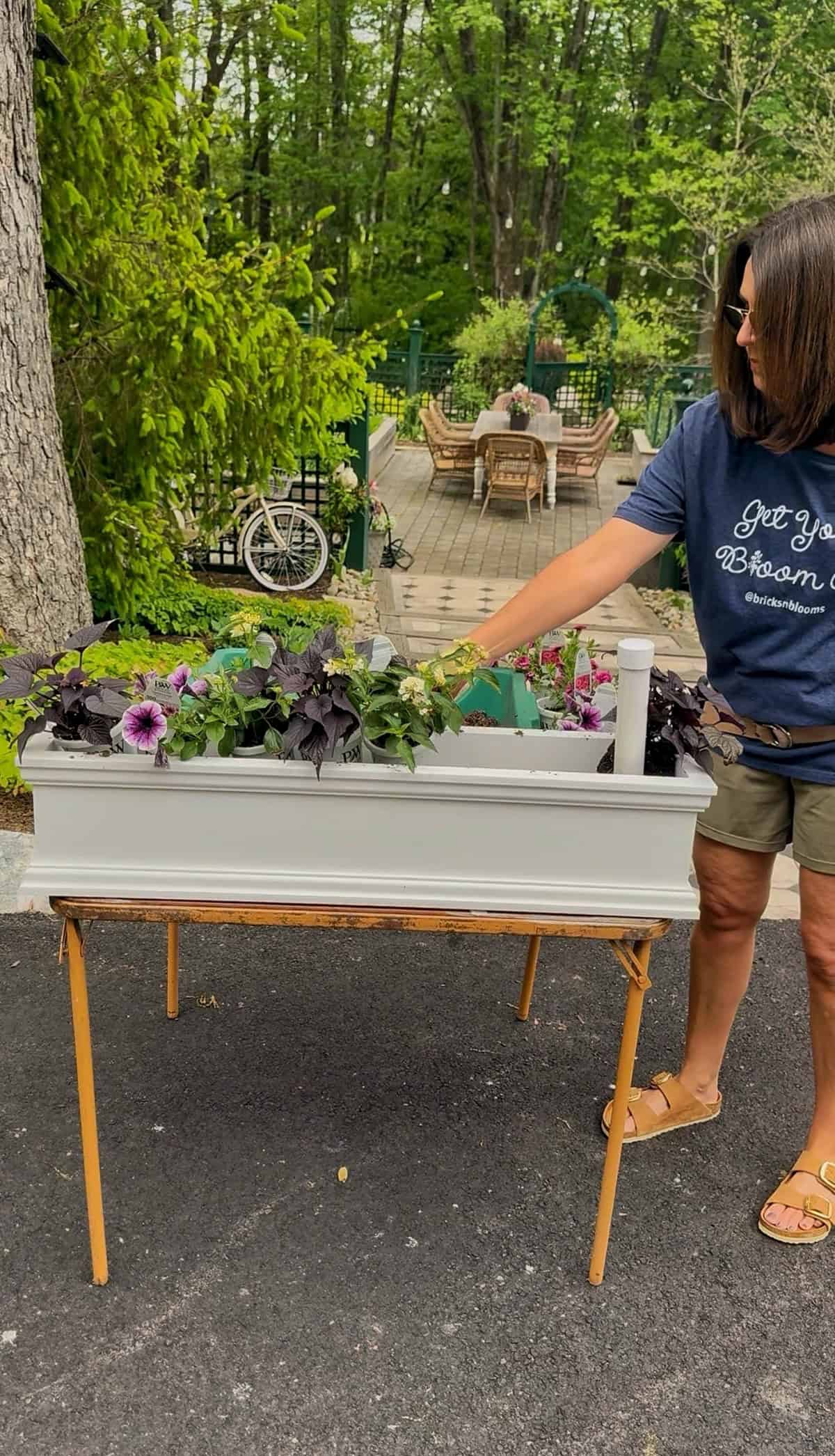 A woman stands beside a raised planter box and nearby window box filled with colorful flowers. She wears a navy t-shirt, shorts, and sandals as she tends the plants outdoors on a patio near a wooded area.