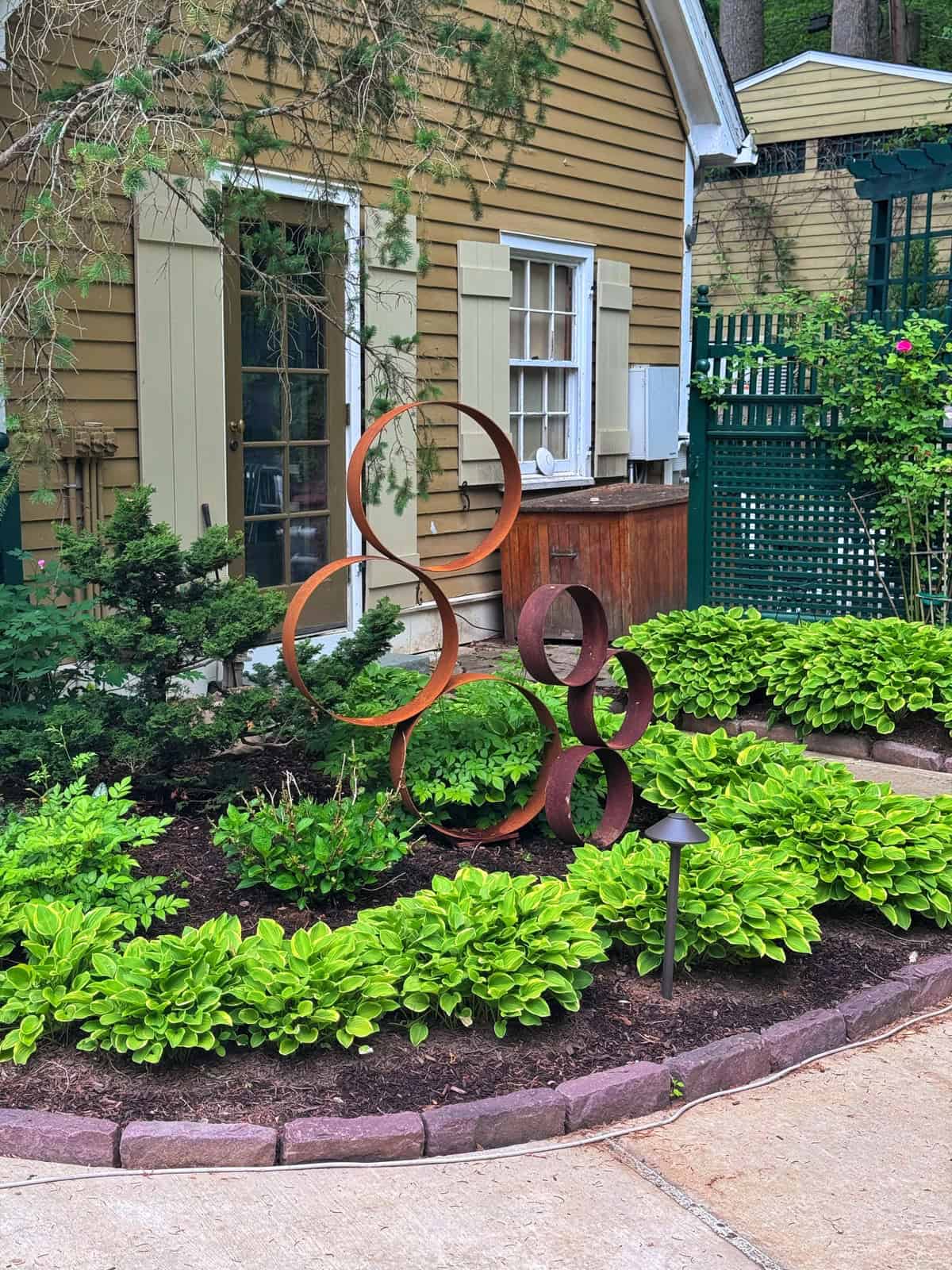 A garden with bright green plants and a modern metal sculpture of interlocking circles in front of a tan house with green shutters and a white window.