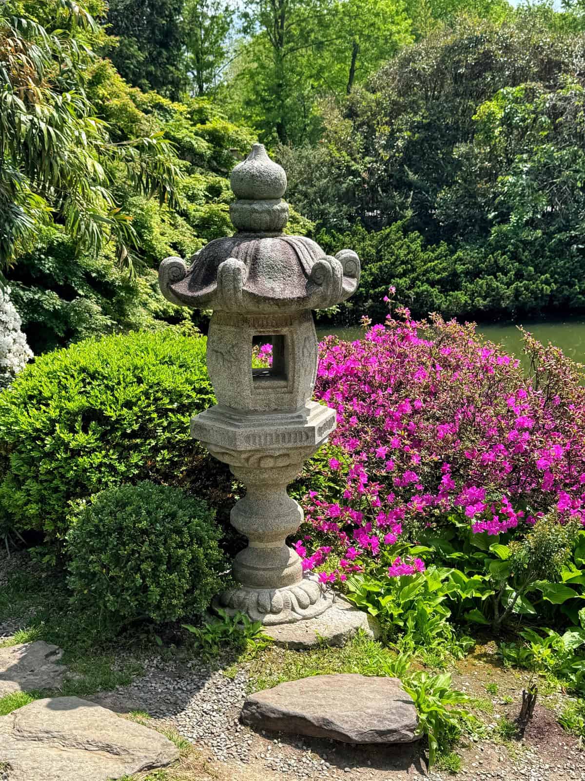 A stone lantern stands in a lush garden surrounded by green shrubs and bright pink flowers, with trees and a pond in the background on a sunny day.