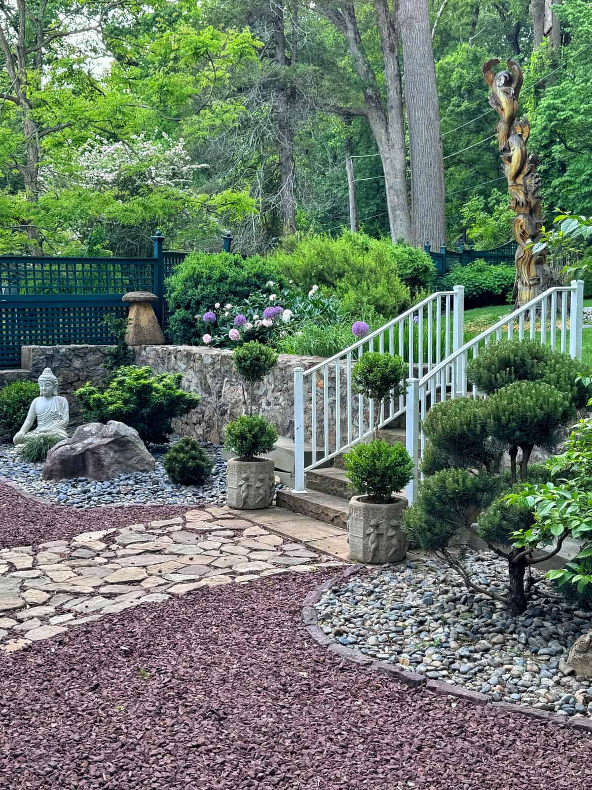 A peaceful garden with a white railing, stone steps, trimmed bushes, a Buddha statue, and a tall wooden sculpture. Pathways of stone and gravel wind through lush greenery and blooming flowers.