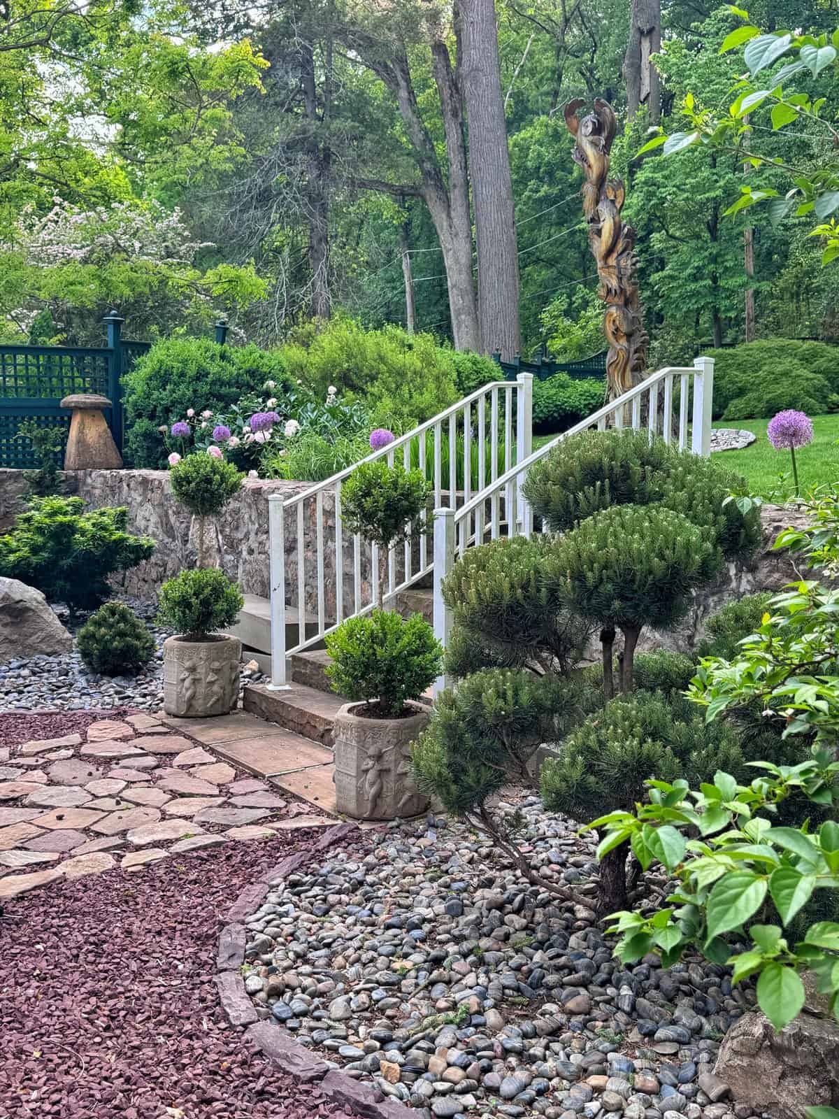 A garden with a white-railed staircase, manicured bushes in stone planters, a stone path, decorative rocks, and lush greenery with trees and flowers in the background.