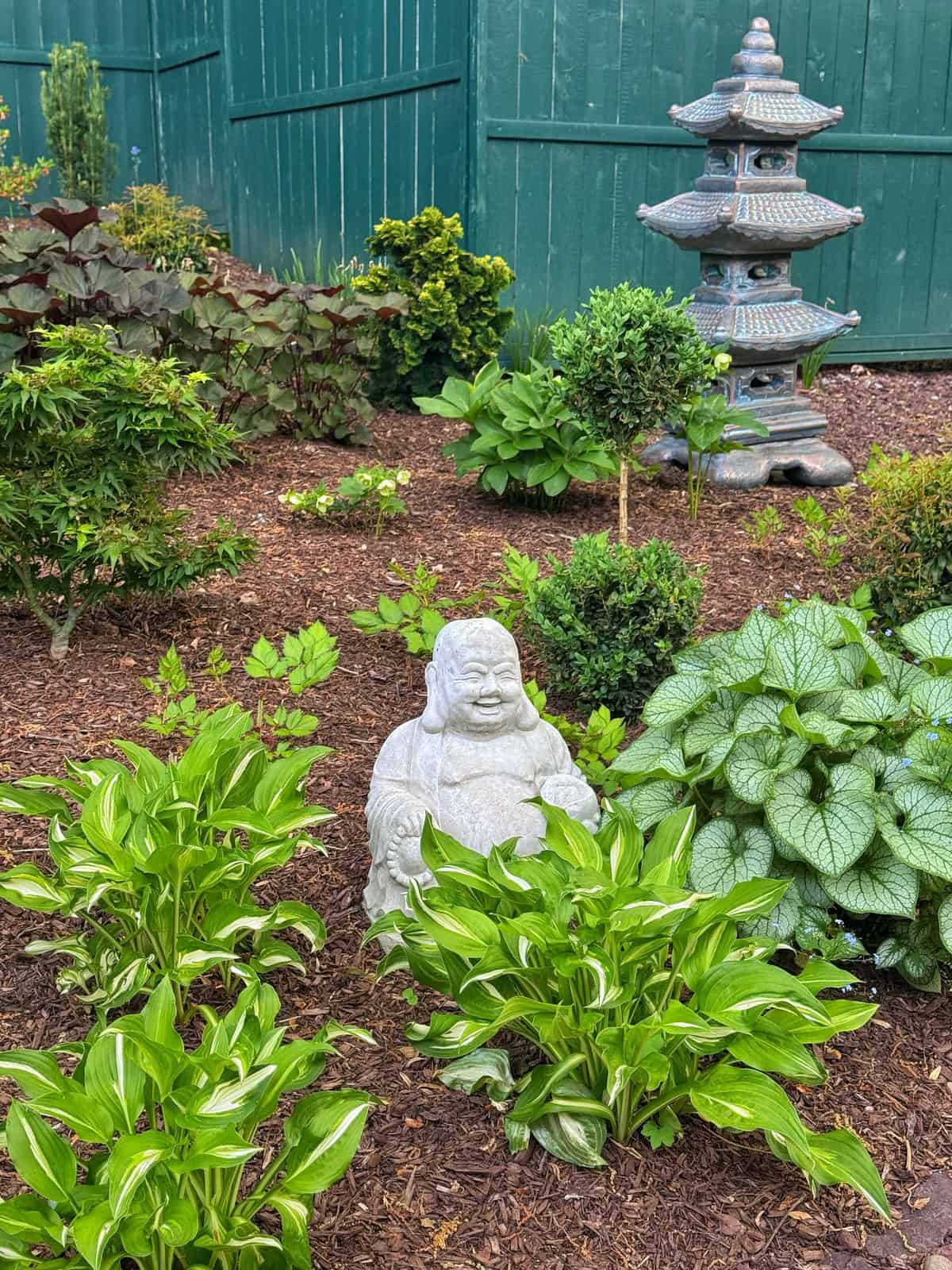 A small stone Buddha statue sits among green plants and mulch in a garden. Nearby, there is a gray pagoda-style garden ornament. A green wooden fence encloses the peaceful scene.