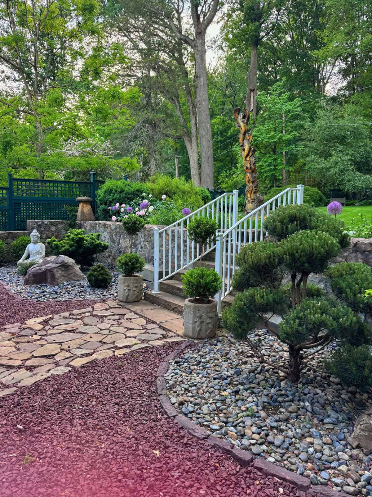A peaceful garden with a stone path, trimmed shrubs, a white railing staircase, potted plants, a Buddha statue, rocks, and green trees in the background. Pink and purple flowers add color to the serene scene.