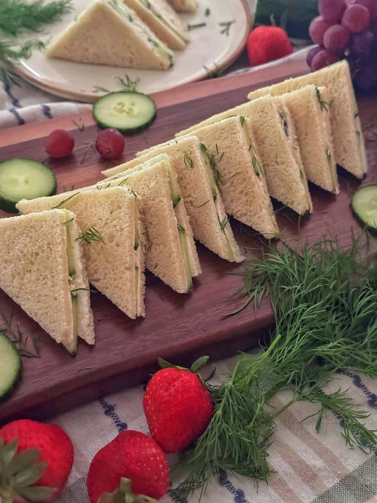 A row of triangular cucumber tea sandwiches on a wooden board, garnished with fresh dill, surrounded by cucumber slices, strawberries, grapes, and more dill. A plate with more sandwiches is in the background.
