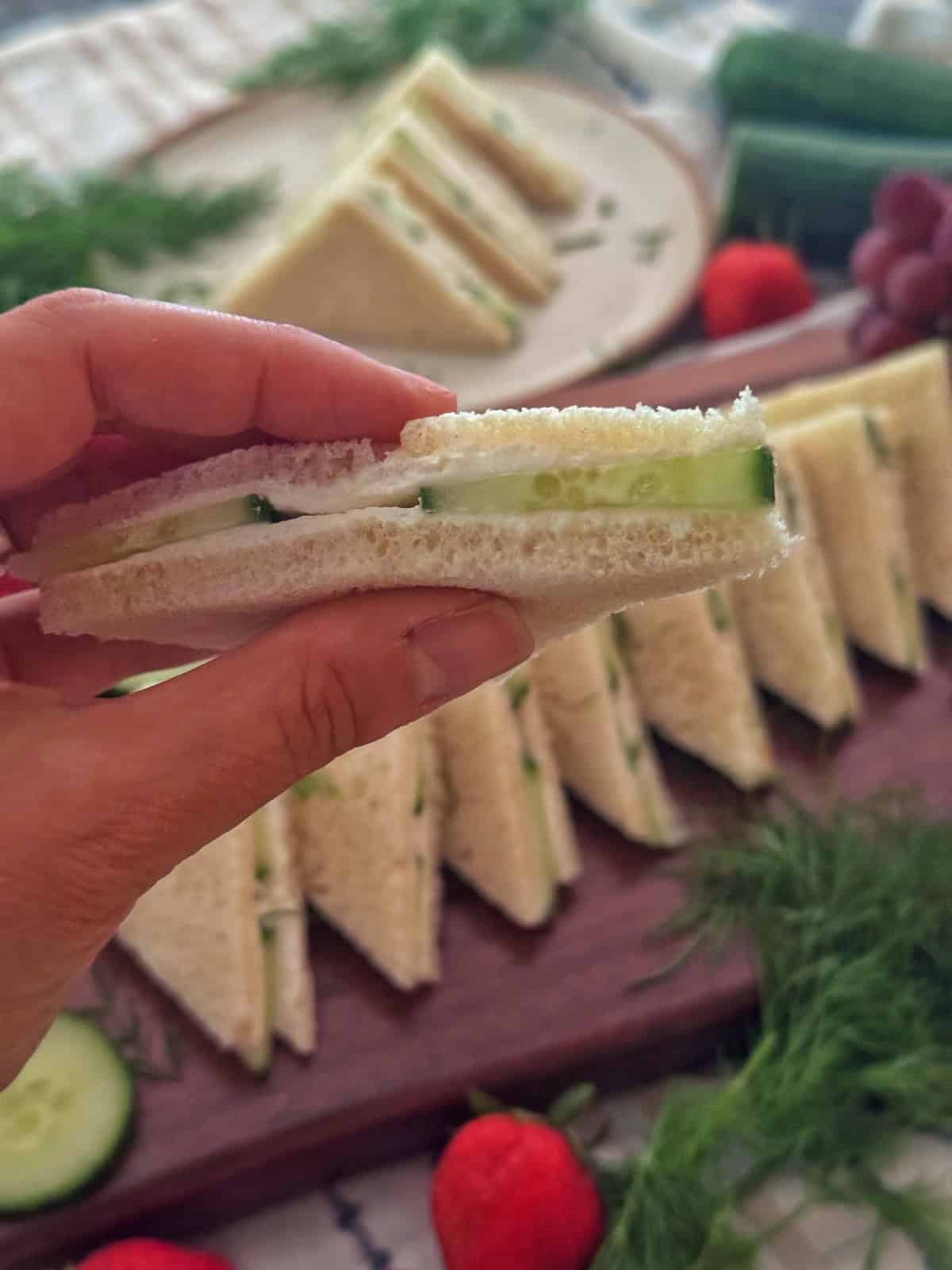 A hand holds a cucumber  cream cheese sandwich with cream cheese on white bread. More sandwiches are neatly arranged on a wooden board with fresh dill, cucumbers, strawberries, and grapes in the background.