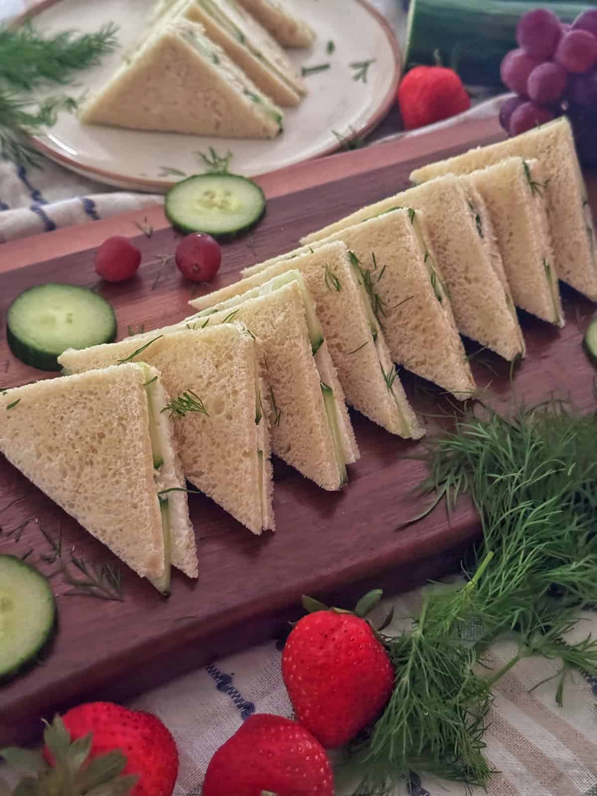 Triangle cucumber cream cheese sandwiches with fresh dill are neatly arranged on a wooden board, surrounded by sliced cucumbers, strawberries, and garnishes. Additional sandwiches and fruit are visible in the background.