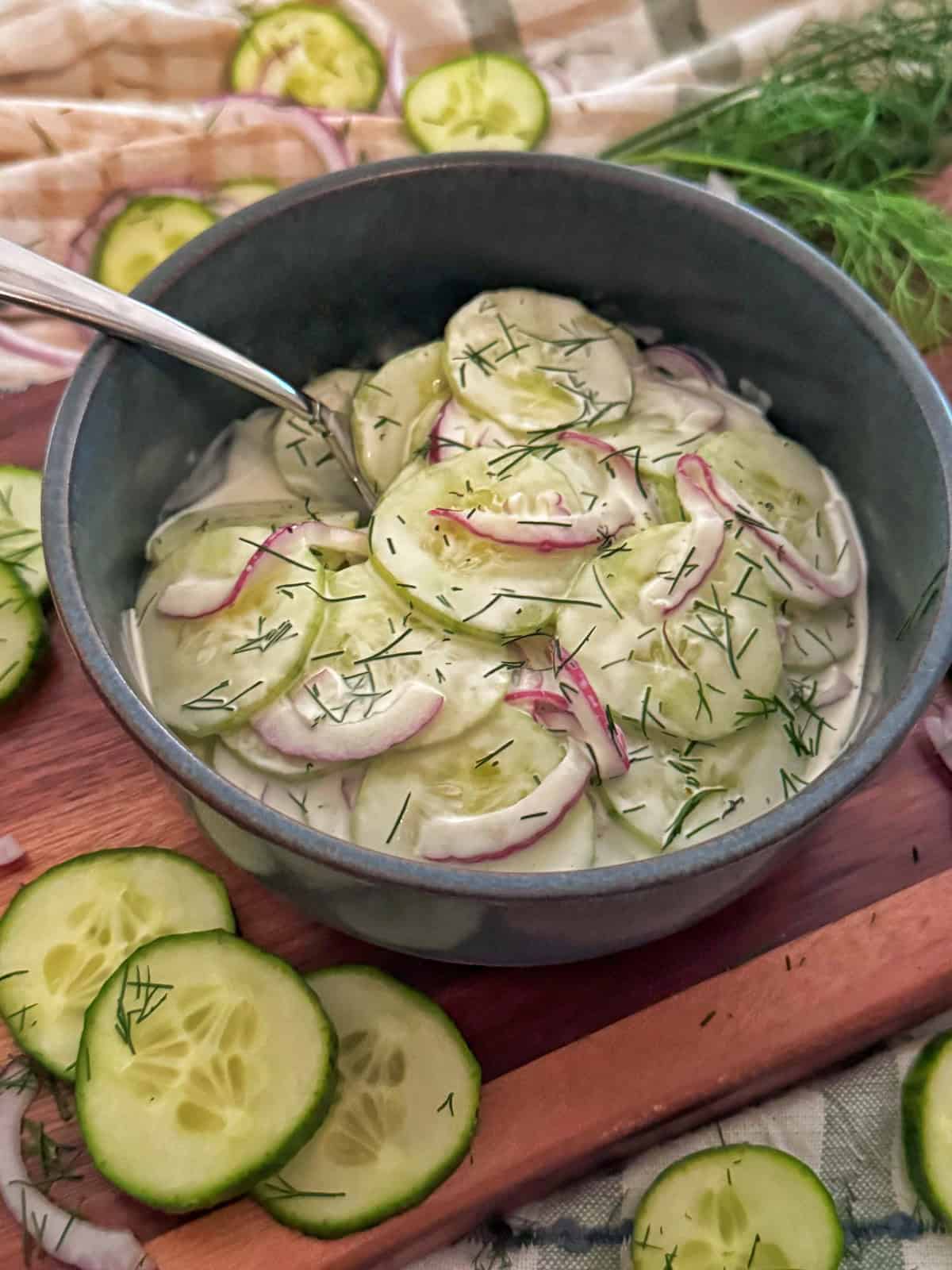 A bowl of creamy cucumber salad with sliced cucumbers, red onions, and fresh dill, garnished with extra dill. Sliced cucumbers and dill are scattered around the bowl on a wooden surface.