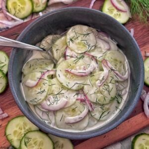 A bowl of creamy cucumber salad with sliced cucumbers, red onions, and chopped dill, served with a spoon. Slices of cucumber, onion, and dill are scattered on the wooden surface around the bowl.