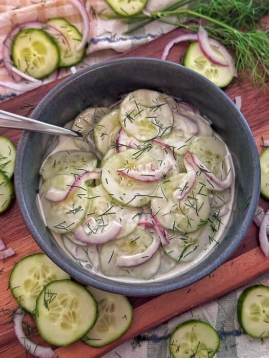 A bowl of creamy cucumber salad with sliced cucumbers, red onions, and chopped dill, served with a spoon. Slices of cucumber, onion, and dill are scattered on the wooden surface around the bowl.