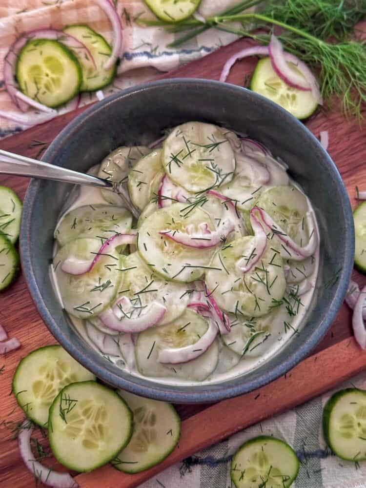 A bowl of creamy cucumber salad with sliced cucumbers, red onions, and chopped dill, served with a spoon. Slices of cucumber, onion, and dill are scattered on the wooden surface around the bowl.