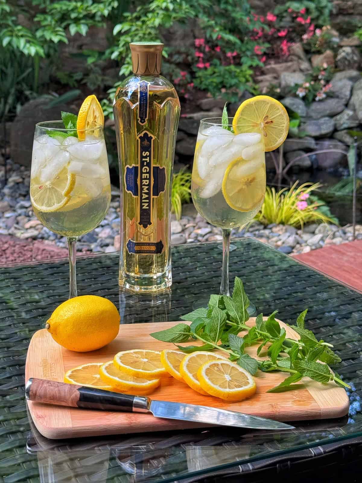 Two glasses of iced cocktails with lemon slices and mint, a bottle of St-Germain, and a cutting board with a knife, lemon slices, and mint leaves on a glass table outdoors, with greenery and rocks in the background.