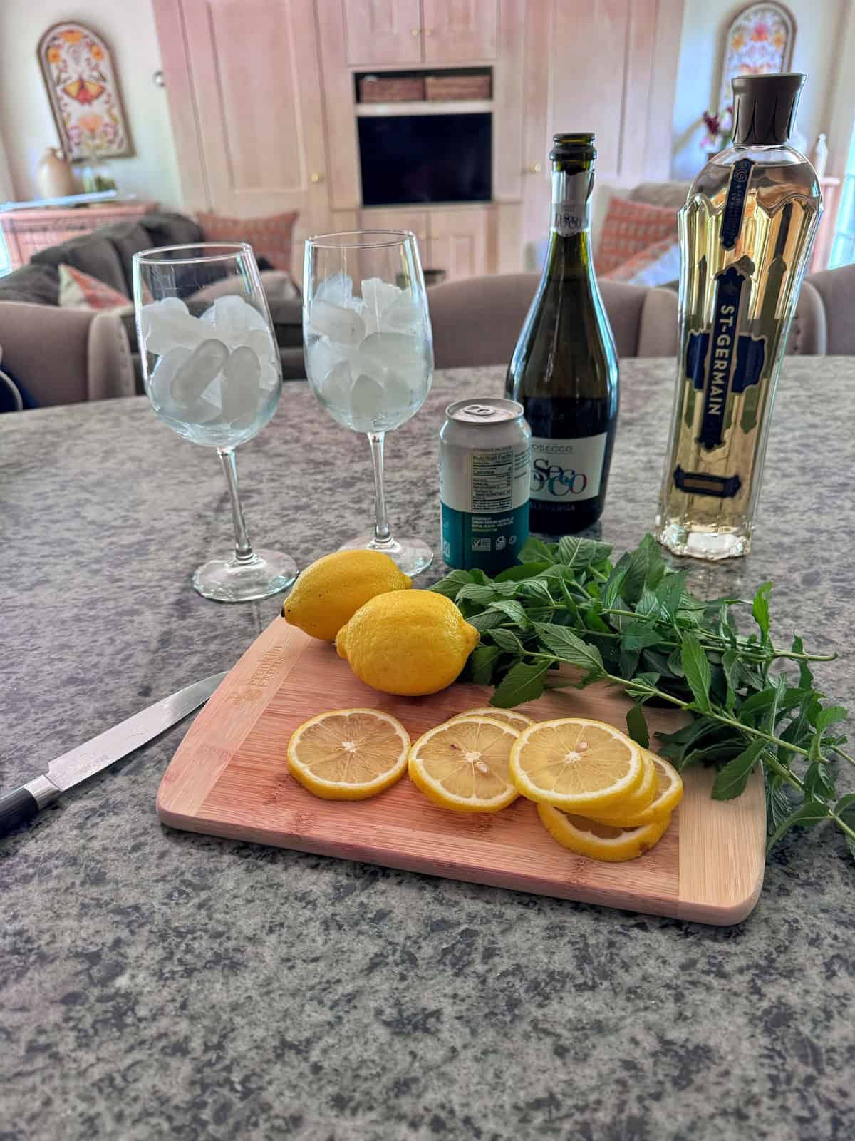 A kitchen counter with two wine glasses filled with ice, a bottle of St-Germain, prosecco, club soda, three lemons (one sliced), fresh mint, and a knife on a cutting board. A cozy living room is in the background.