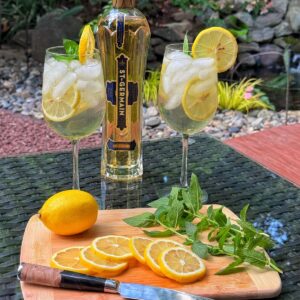 A bottle of St-Germain liqueur stands behind two iced hugo spritz cocktails garnished with lemon and mint on a glass table. In front, a wooden board holds a knife, lemon slices, a whole lemon, and mint sprigs. Lush garden in background.