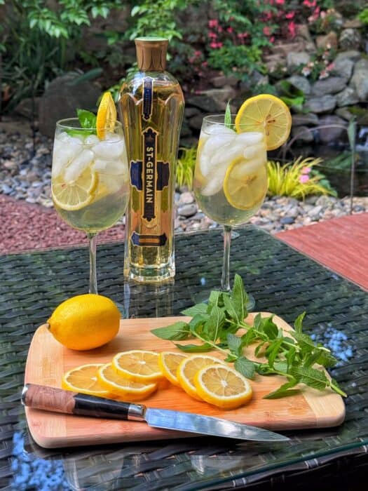 A bottle of St-Germain liqueur stands behind two iced hugo spritz cocktails garnished with lemon and mint on a glass table. In front, a wooden board holds a knife, lemon slices, a whole lemon, and mint sprigs. Lush garden in background.