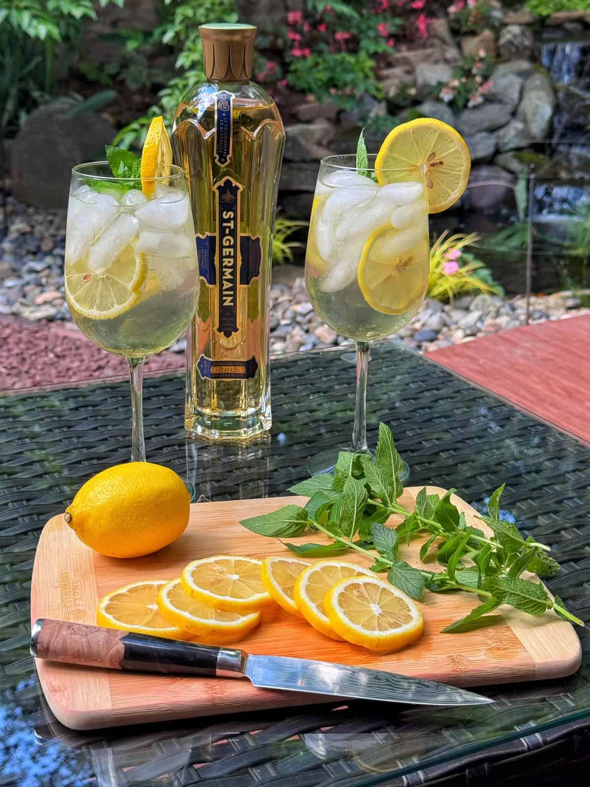 Two glasses with ice, lemon slices, and mint sit on a table next to a St-Germain bottle. A wooden board holds a knife, sliced lemon, a whole lemon, and fresh herbs. A garden with rocks and plants is in the background.