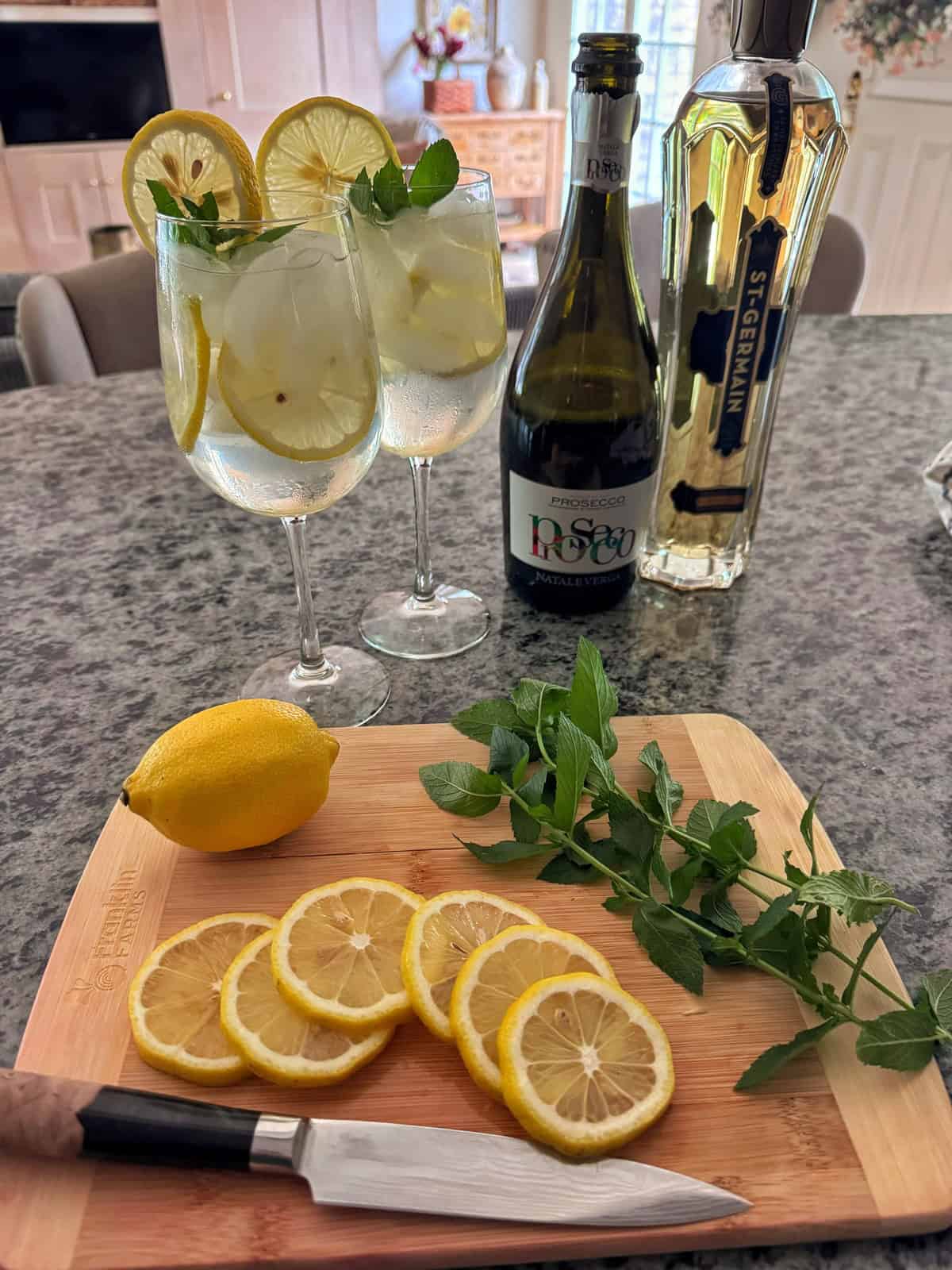 Two glasses of a lemon and mint cocktail with ice, beside a bottle of St-Germain liqueur and a bottle of sparkling wine. A cutting board holds sliced lemon, a whole lemon, mint sprigs, and a knife.