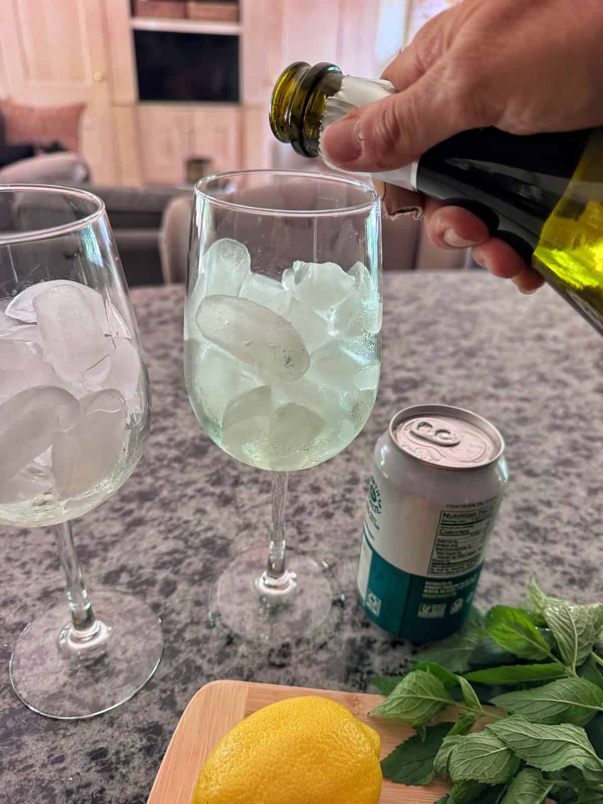 A hand pours a prosecco into a wine glass filled with ice. Another glass with ice, a can, lemon, mint leaves, and a wooden tray are on a gray countertop in a cozy living room setting.