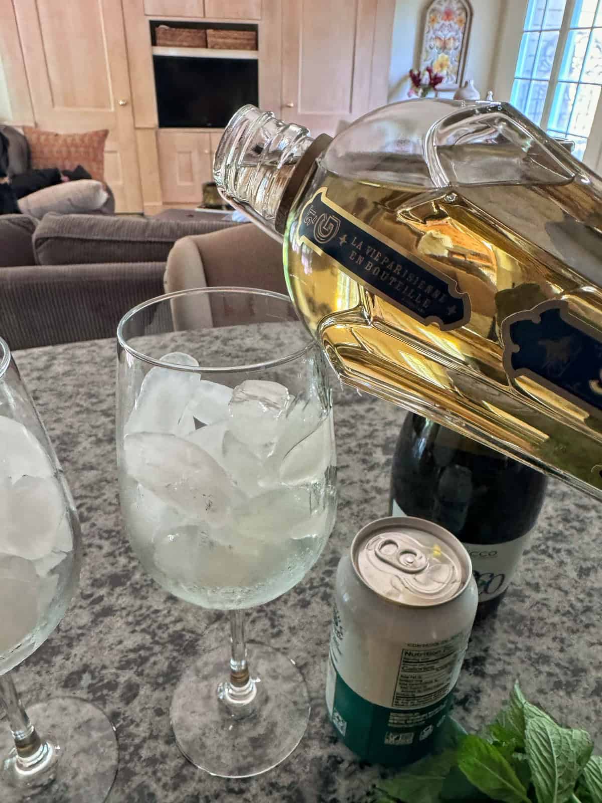 A close-up of a bottle of st germain
 being poured over ice in a wine glass. Next to the glass are another glass with ice, a can of club soda, a bottle of prosecco, and some fresh mint on a countertop.