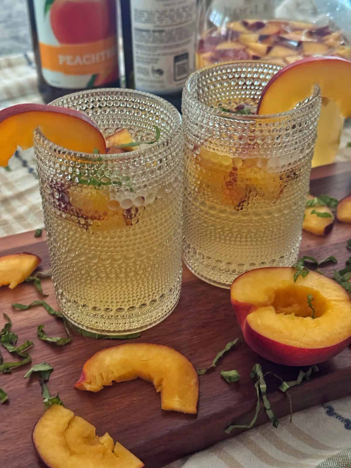 Two clear textured glasses filled with peach sangria, garnished with fresh peach slices and herbs, sit on a wooden board with scattered peach slices and chopped herbs. Bottles are blurred in the background.