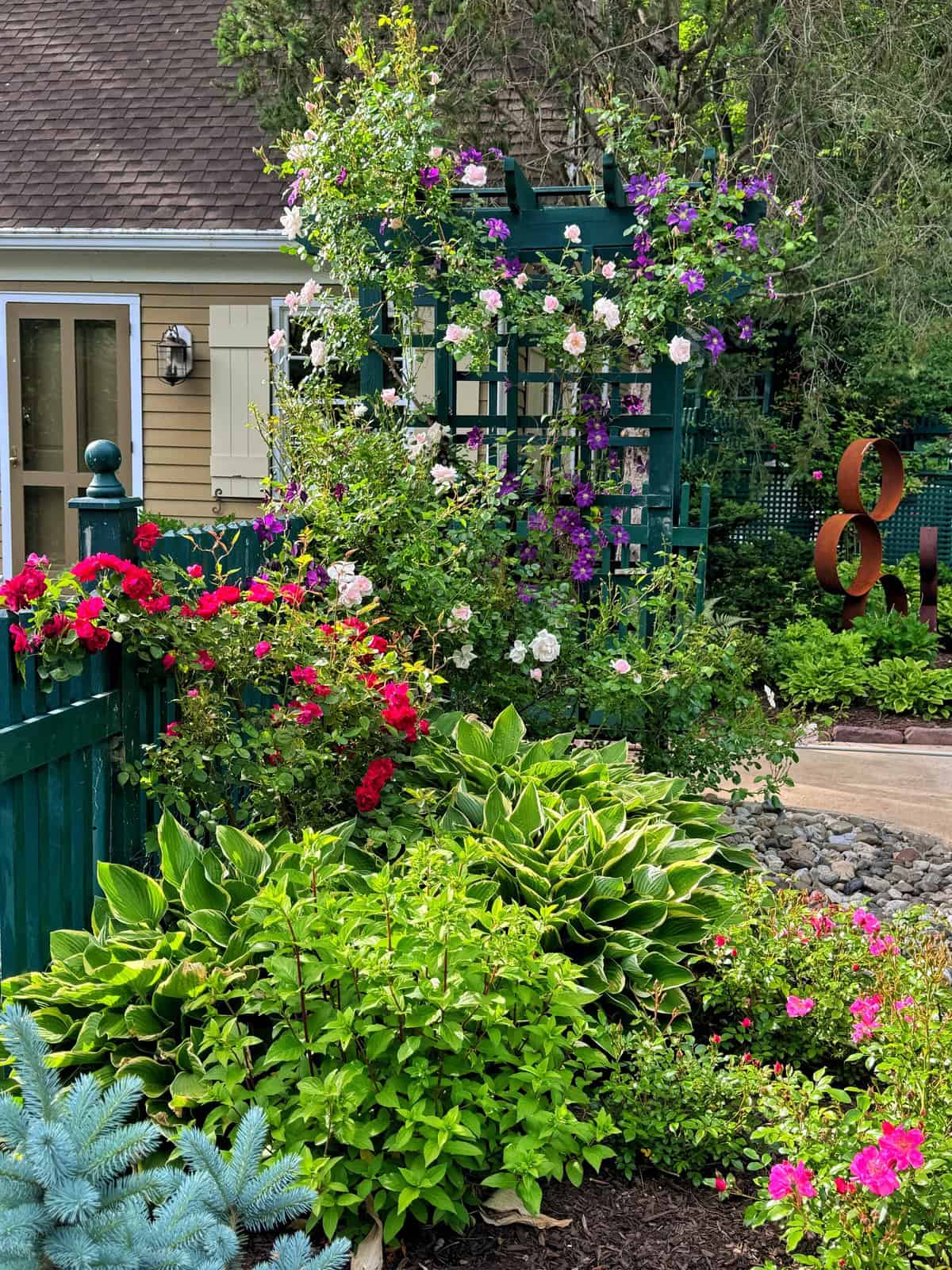 A lush garden with blooming red, pink, and purple flowers, green shrubs, and leafy plants surrounds a green trellis and fence in front of a yellow house. A large metal &ldquo;8&rdquo; sculpture is visible in the background.
