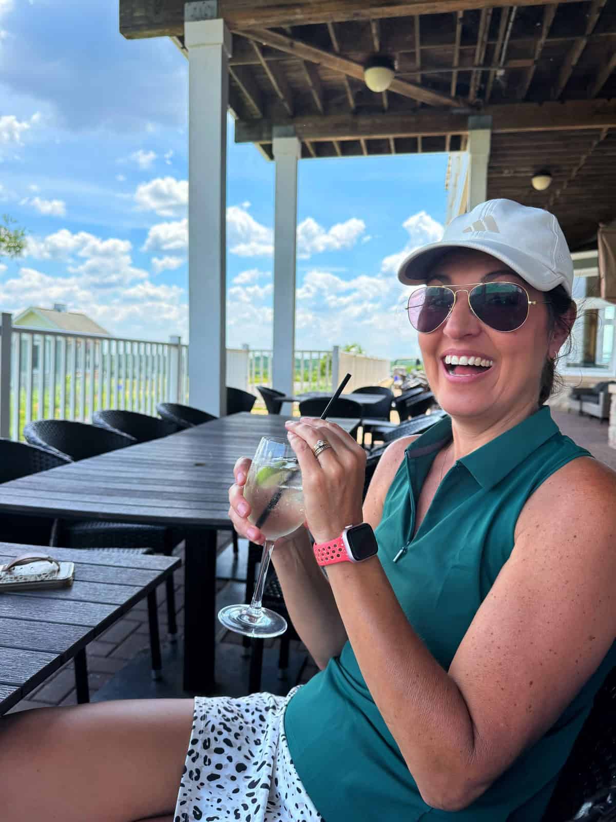 A woman wearing sunglasses, a white cap, and a sleeveless green shirt smiles while holding a drink on an outdoor patio under a covered roof. The day is sunny, with a blue sky and clouds visible in the background.