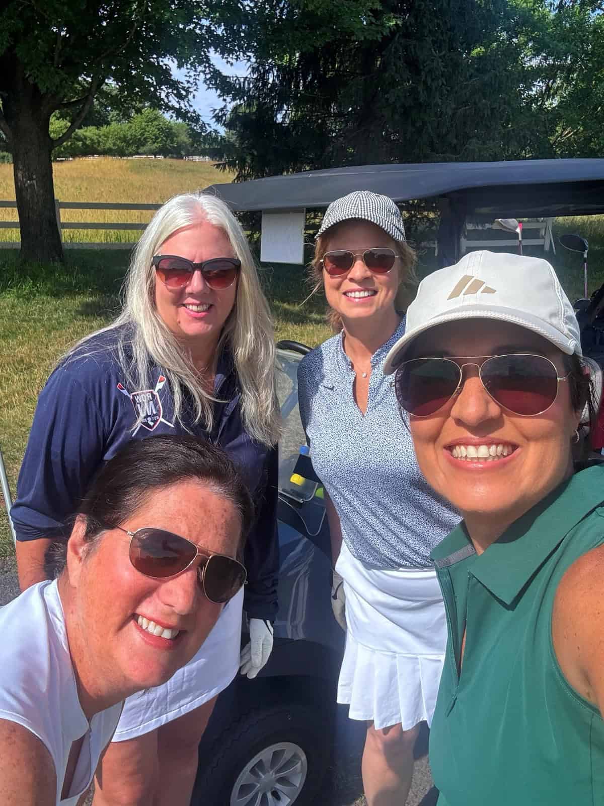 Four women smiling and posing for a selfie outdoors on a sunny day, standing in front of a golf cart, dressed in golf attire with trees and a grassy area in the background.