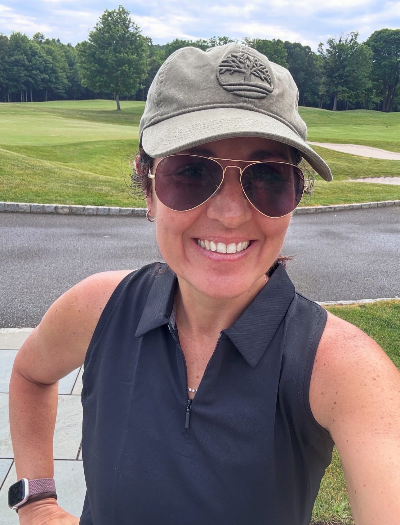 A woman (stacy ling) wearing aviator sunglasses, a khaki cap, and a sleeveless black top smiles outdoors on a golf course with green grass and trees in the background.